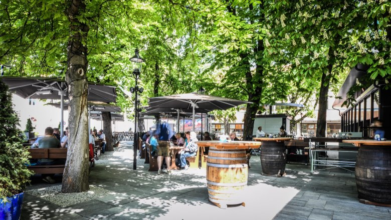 Hofbräu, © Extra Krems A beer garden with wooden tables and barrels under green trees, people sit and enjoy the atmosphere.