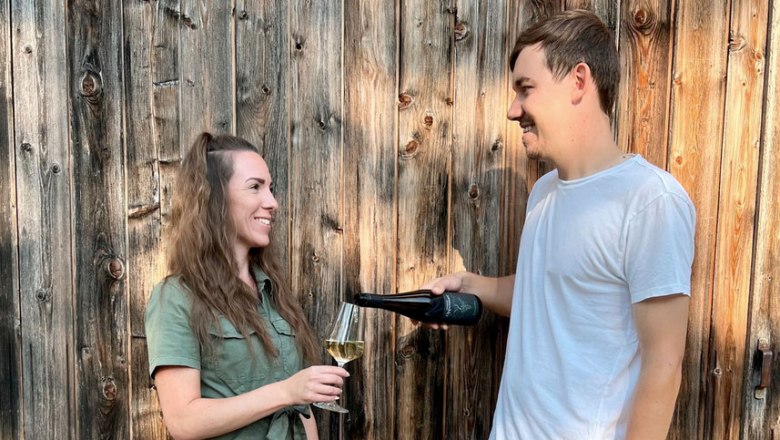 Rainer and Lisa, © Bioweingut Oppenauer A man pours wine for a woman, both smiling in front of a wooden wall.