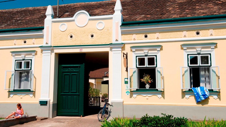 Winzerhof & Guesthouse Greil, © M. Greil A traditional yellow building with green doors and windows, a bicycle in front of it and a child sitting on the stairs.