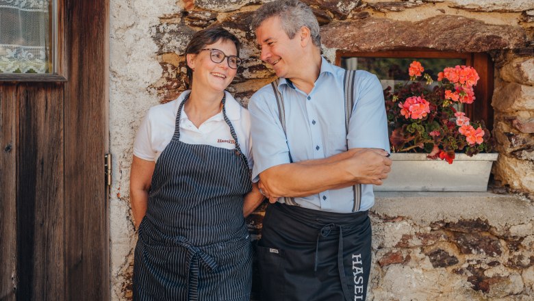 Waltraud & Paul Haselböck, innkeepers, © Niederösterreich Werbung/Daniela Führer A man and a woman in aprons lean smiling against a stone wall next to a window with flowers.