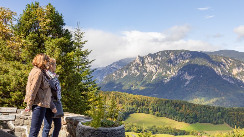 Impressive view from the Looshaus, © Wiener Alpen / Christian Kremsl Two people stand on a terrace and look out over a mountain landscape with forests and meadows.