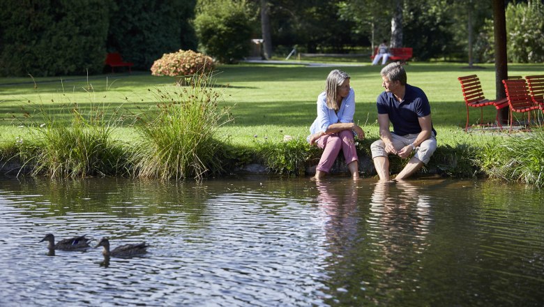 Bad Schönau spa gardens, © Wiener Alpen, Florian Lierzer A couple sits on the bank of a pond in the spa gardens of Bad Schönau, their feet in the water. Two ducks swim in the foreground.