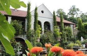 Nursery-Atzenbrugg, © Lederleitner A white building with a red tiled roof, surrounded by lush vegetation and orange flowers in the foreground.