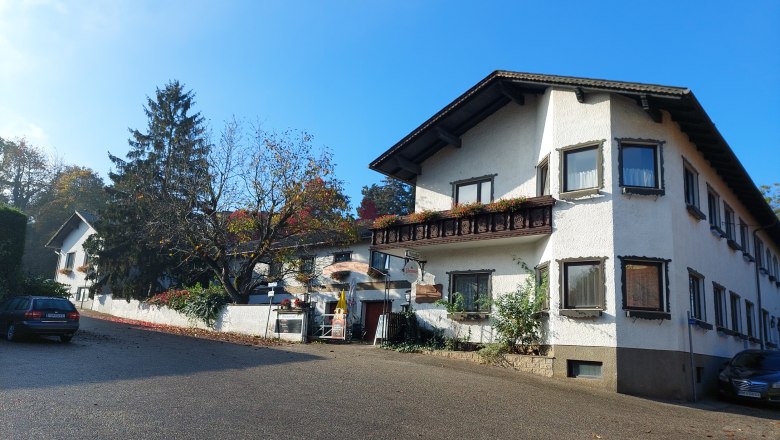 TDR-Welserhof-outside, © Nora Bartfai Exterior view of a traditional building with white walls and wooden decorations, surrounded by trees and a blue sky.