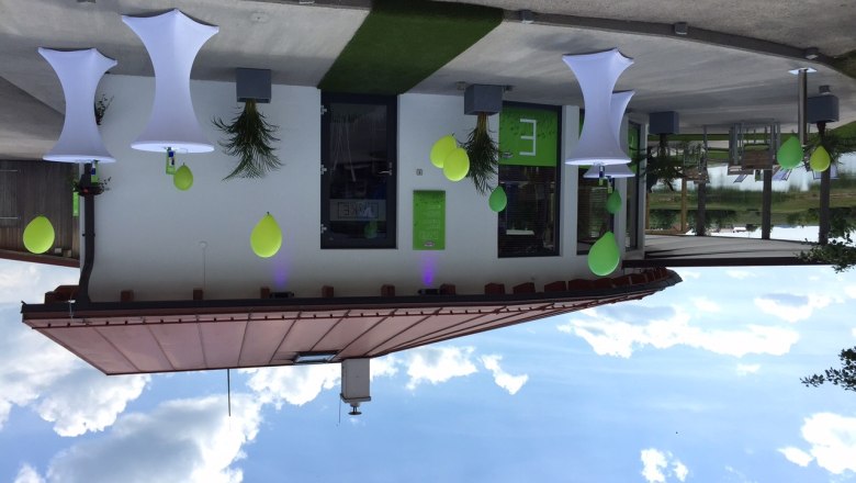 Friedrich beach bar, © Gemeinde Jaidhof A modern beach bar with white bar tables and green decorations in front of a building with a flat roof.