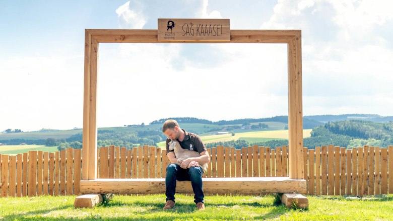 Mandl's goat farm, © Sooo gut schmeckt die Bucklige Welt/Viktoria Kornfeld A man sits on a wooden bench under a large frame with the inscription 'SAG KÄÄÄSE' and holds a kid. A rural landscape can be seen in the background.