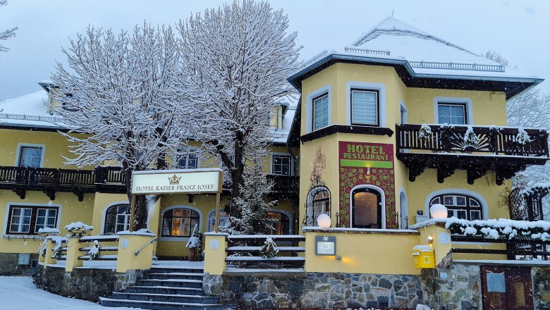 Winter - Hotel Kaiser Franz Josef, © Matthias Strutzmann A yellow hotel with the name Kaiser Franz Josef in winter, surrounded by snow-covered trees and a snowy forecourt.