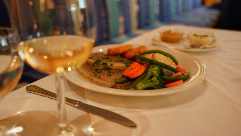 Culinary delights, © Hotel Stich A plate of meat, vegetables and potatoes on a table, with a glass of white wine next to it.