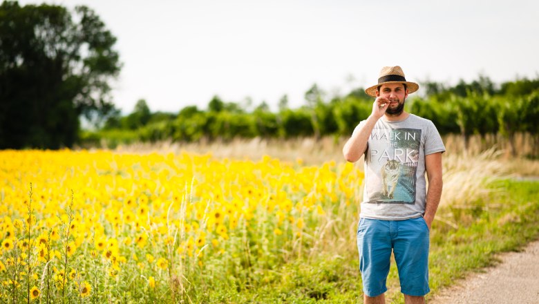 Martin Obenaus, © Bernhard Kastner Man in a straw hat stands on a path next to a field of sunflowers.