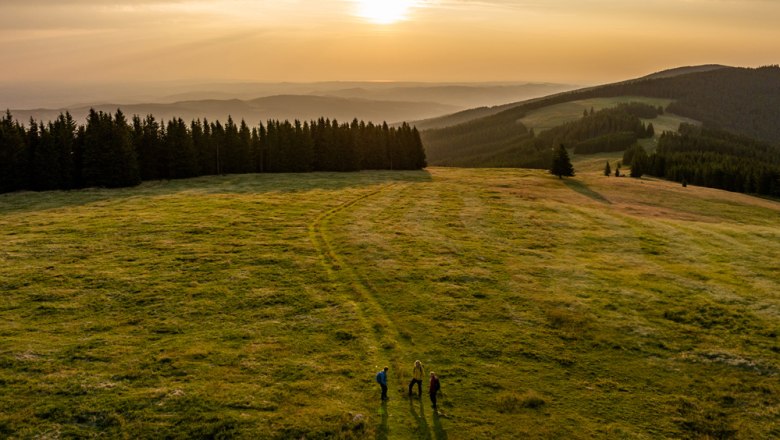 Morning mood near the Feistritzer Schwaig, © Wiener Alpen, Kremsl Three people walk across a green meadow at sunrise, surrounded by woods and hills.