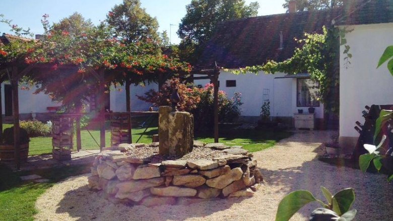Garden, © Pollak A sunny garden with a stone circle and a small fountain in the middle, surrounded by white buildings and green plants.