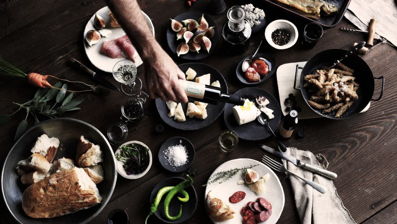 Gilli Mill - Tastings, © K. Vyhnalek A rustic table with various dishes, including bread, cheese, figs, sausage and fish. A hand pours oil over a dish.
