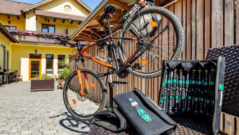 Cycling host Hotel Post-Hönigwirt, © Wiener Alpen/Martin Fülöp An orange bicycle on a stand in front of a yellow building with a wooden fence and tool set in the foreground.