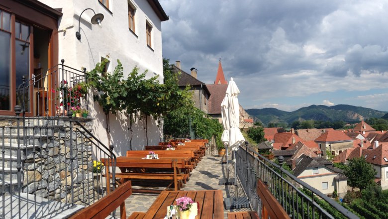 Outlook, © Turm Wachau Terrace with wooden benches and tables, view of rooftops and mountains in the background.