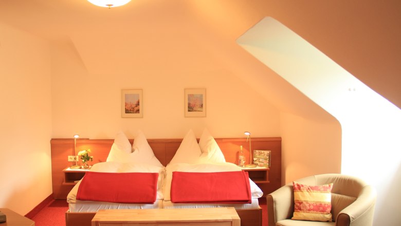 Double room Classic, © Schendl A cozy double room with two beds, red blankets, an armchair and a table under a sloping roof.