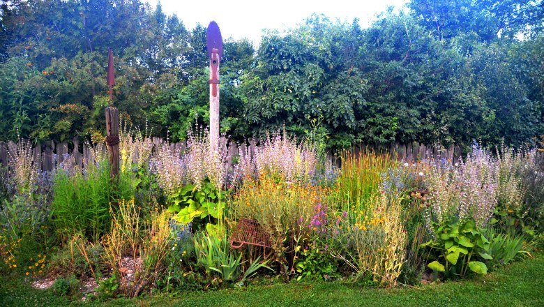 perennial_bed_small_contrast_c_family-mahr, © Familie Mahr A colorful perennial bed with various flowers and plants in front of a wooden fence and trees.