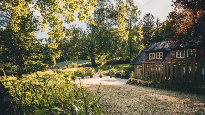 Country estate Oberhof, © Niederösterreich Werbung / Maximilian Pawlikowsky A rural garden with a small house and a gravel path, surrounded by trees and plants in the sunlight.