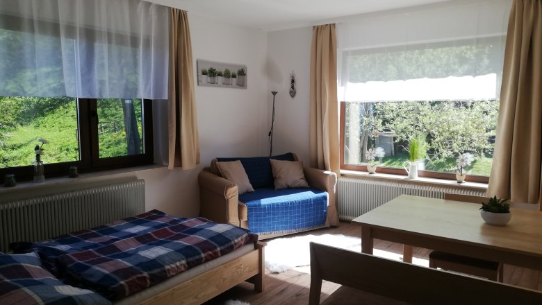 Vacation apartment, © Familie Weibels Bright living room with bed, sofa, table and large windows with a view of the greenery.