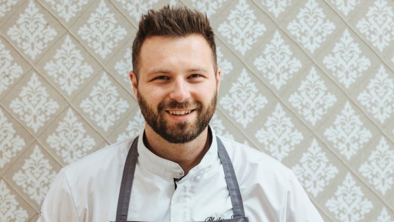 Chef and host Philipp Essl, © Niederösterreich Werbung/Michael Reidinger Philipp Essl in chef's clothes in front of a patterned wallpaper.