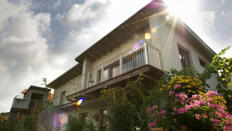 Wooden hay exterior view, © Philipp Monihart Exterior view of a house with balcony and flowering garden in the foreground, rays of sunshine in the sky.