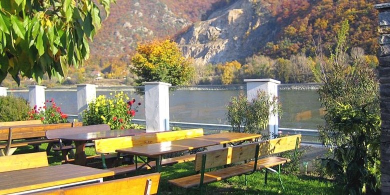 Wine tavern, © Weingut Wagner A wine tavern with wooden benches and tables on the riverbank, surrounded by autumnal trees.