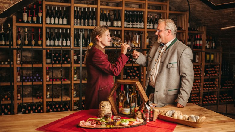 Wine tastings in the atmospheric wine cellar, © Niederösterreich Werbung / Daniela Führer Two people clink glasses in a wine cellar, surrounded by bottles of wine and a table with snacks.