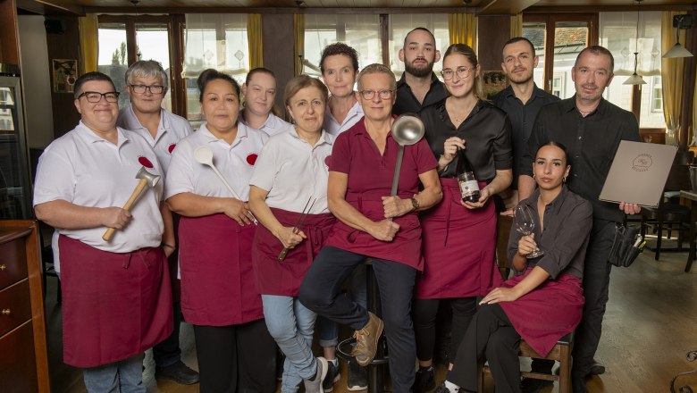 Team around Birgit Reisinger, © Foto Buttinger A team of restaurant staff pose in work clothes with kitchen utensils in a dining room.