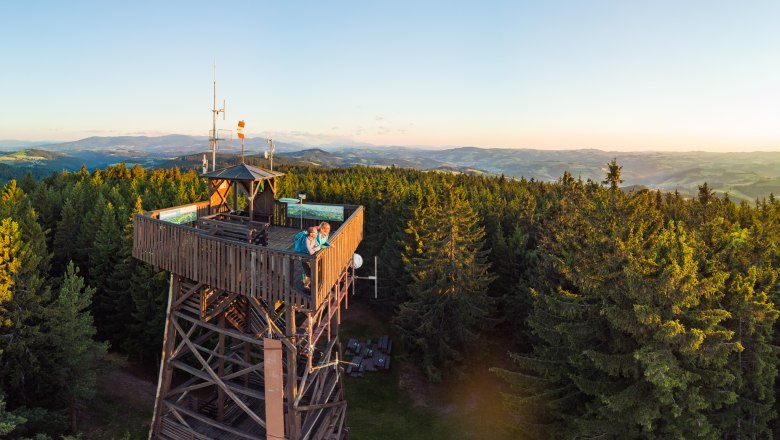 The lookout point at Hutwisch, © Wiener Alpen/Martin Fülöp Wooden observation tower in the forest with two people watching the sunset, hilly panorama in the background,