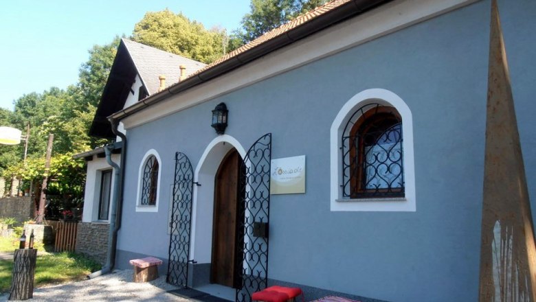 Loessiade, © Lössiade A small, blue building with a wooden door and decorative bars in front of the windows. In front of it are wooden stools with red cushions.