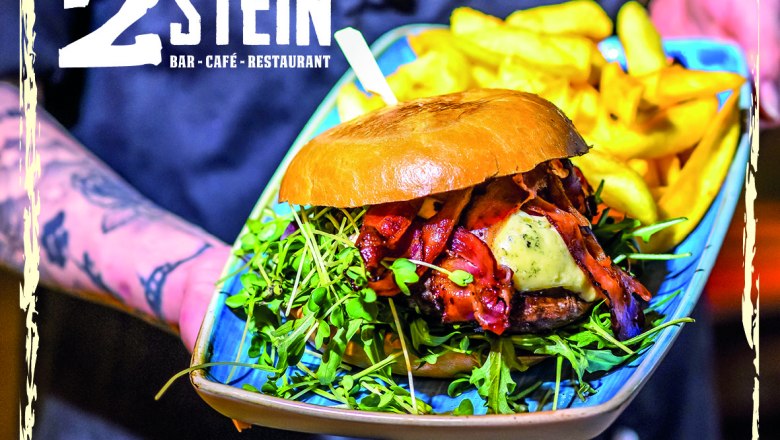 2Stein Bar-Café-Restaurant, © 2Stein A burger with bacon, cheese and rocket on a plate with fries, presented by a tattooed hand. Logo of 2Stein Bar-Café-Restaurant top left.