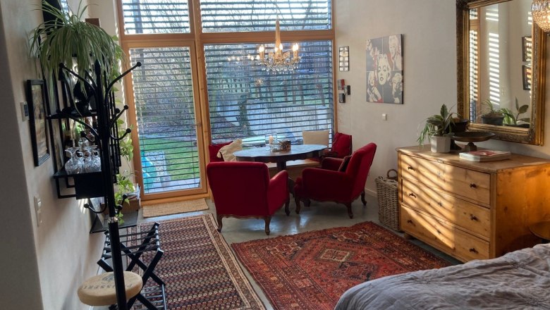 Apartment with a view of the garden, © Weitschacher Living room with large windows, red armchairs, wooden furniture and carpets, view of the garden.