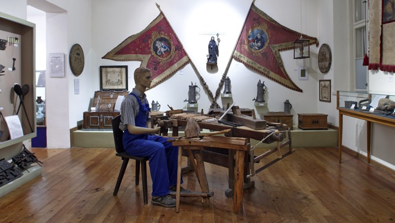 Neunkirchen Municipal Museum, © Wiener Alpen/Bene Croy Interior view of a museum with a mannequin in work clothes at a wooden table, surrounded by historical objects and flags.