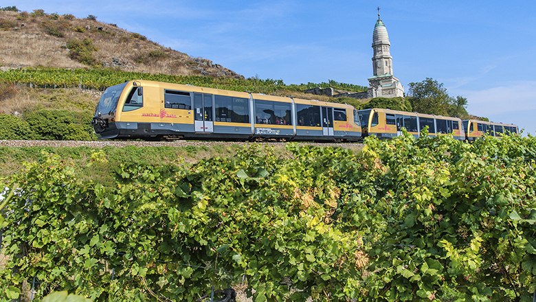 Wachau Railway, © NB/Kerschbaummayr A yellow Wachau Railway train travels through a green vineyard landscape with a church in the background.