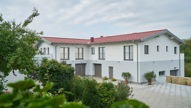Weinhof Maier, © Weinhof Maier A modern building with a red roof and white walls, surrounded by green trees and plants.