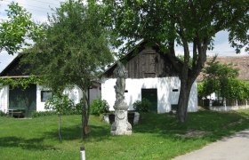 Wine cellar lane Großkadolz, © Gemeinde Seefeld-Kadolz A white building with a wooden gate and statue in the foreground, surrounded by trees and green lawns.