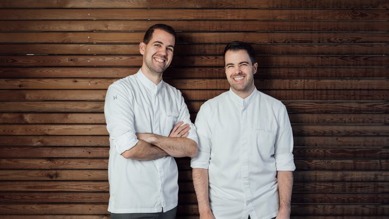 The brothers: Martin and Stefan Görg, © Wiener Alpen, Sophie Menegaldo Two men in white shirts stand in front of a wooden wall and smile.