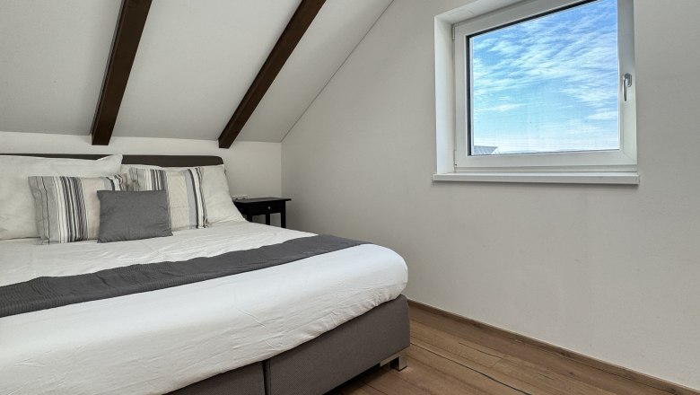 Bedroom, © Juwel am Stadtrand Modern bedroom with wooden beamed ceiling, large bed and window with a view of the blue sky.
