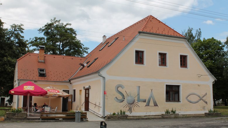 SOLA Safov, © Hannes Messmann A yellow building with a red tiled roof and the word 'SOLA' on the wall. There are parasols and tables in front of the building.