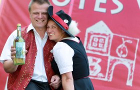 Zarl family, © weinfranz.at A man and woman in traditional dress smile and hold a bottle of apple and pear cider against a red background.