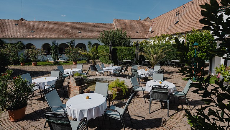 Relax in the cozy Garaten, © Haimer/Michael Reidinger A cozy garden with round tables and chairs, surrounded by plants and an old building with a tiled roof.