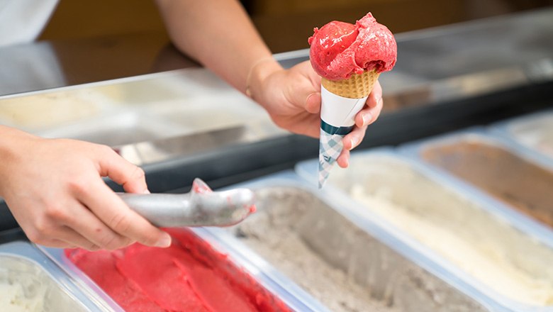 One hand holds an ice cream cone with a scoop of fruit ice cream, the other holds an ice cream scoop., © Blochberger Eisproduktion GmbH One hand holds an ice cream cone with a scoop of fruit ice cream, the other holds an ice cream scoop.