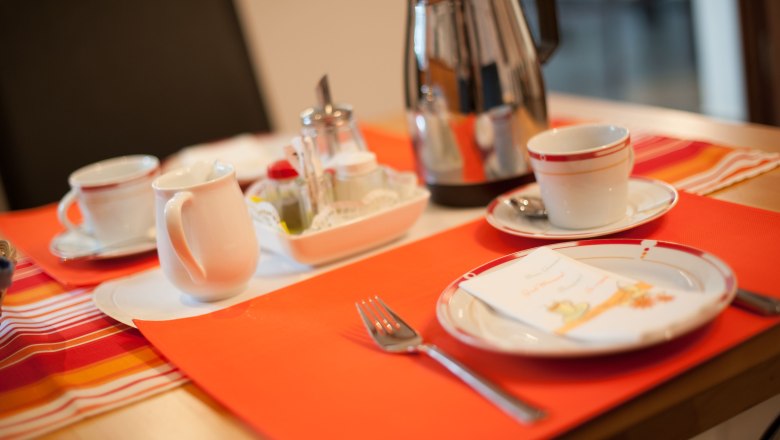 Breakfast, © Fam. Edlinger Breakfast table with cups, plates, cutlery and coffee pot on an orange place mat.