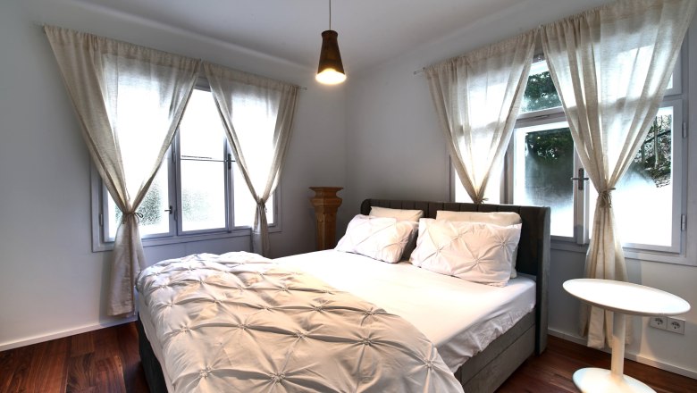 Bedroom Top 2, © Daniela Sinadinoska A cozy bedroom with a large bed, white curtains and a small round table.