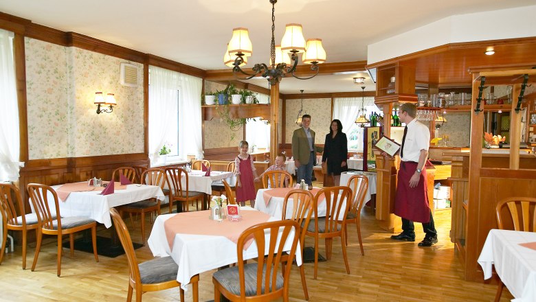 Rustic charm in the living room, © Hausmann/Zwickl A rustic restaurant with wooden furniture and tablecloths. A waiter greets a family at the entrance.