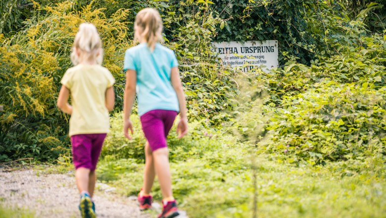 In the Leitha-Au, © Wiener Alpen, Martin Fülöp Two children are walking along a path in a green wooded area. An ivy-covered sign with the inscription 'Leitha Ursprung' is visible in the background.