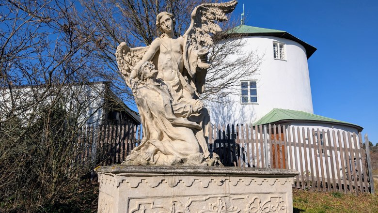 Calvary, © Retzer Land / Daniel Wöhrer Stone statue of an angel with wings and a kneeling figure in front of a round, white building with a green roof.
