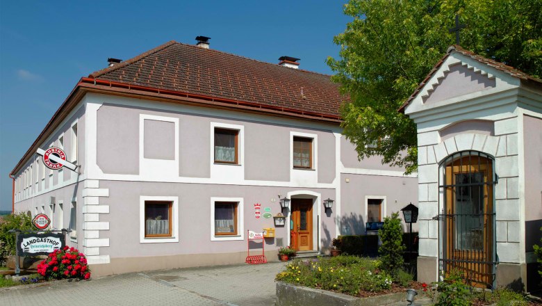 Country inn Heinrichsberg, © Martin Brack, Kilb zVg A traditional country inn with a gray façade and red roof tiles, surrounded by flowers and trees.