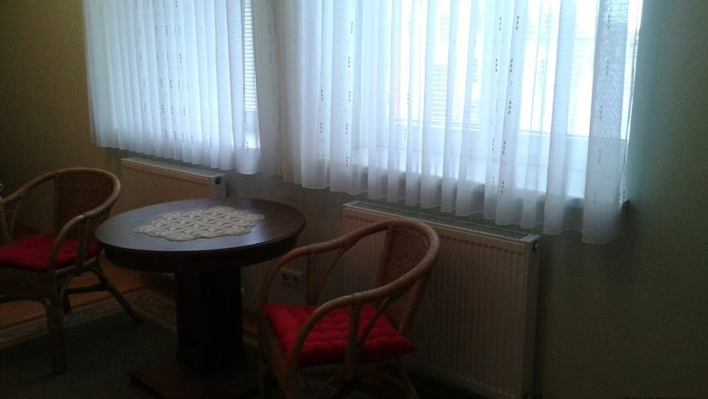 Seating in the triple room, Waysmayer family, © © Bianca Waysmayer Two wicker chairs with red cushions and a round wooden table in front of a window with white curtains.