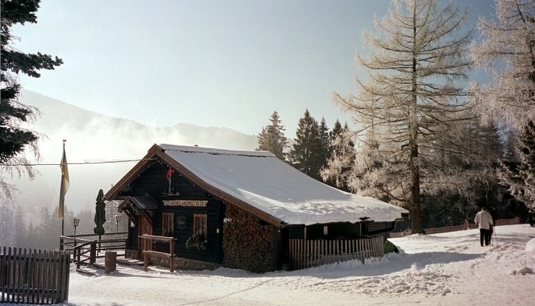 Grenzland hut, © Grenzlandhütte, Foto Paul Laschitz A snow-covered mountain hut in a wintry landscape with trees and a walker.