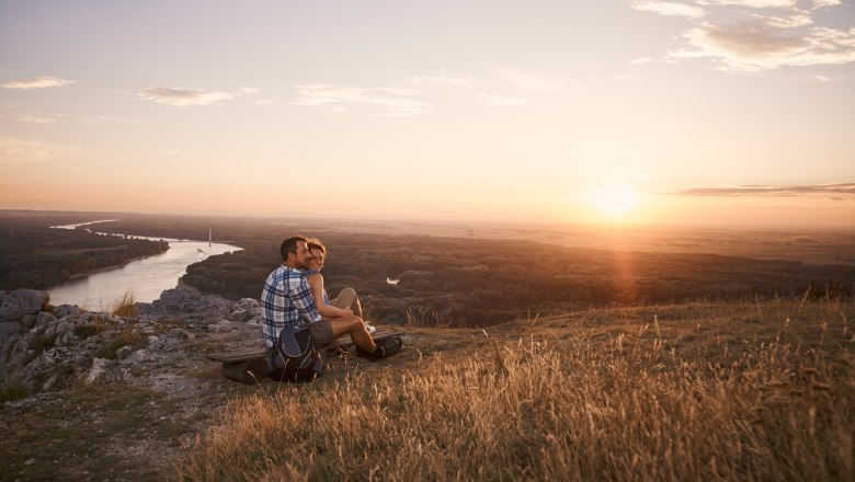 Braunsberg, VIA.CARNUNTUM., © Donau Niederösterreich, Andreas Hofer A couple sits on a hill and looks out over a river and the landscape at sunset.